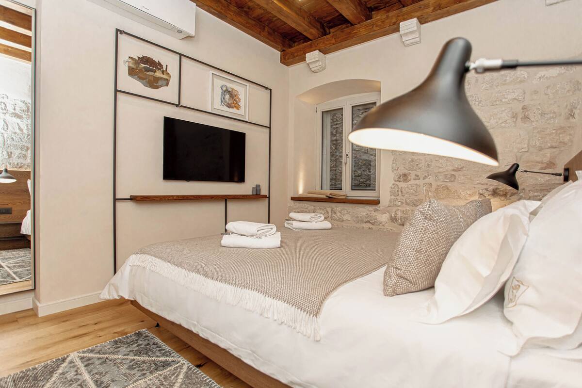 A bedroom with a bed, a TV mounted on the wall, a window with a view, and a lamp on the bedside table.