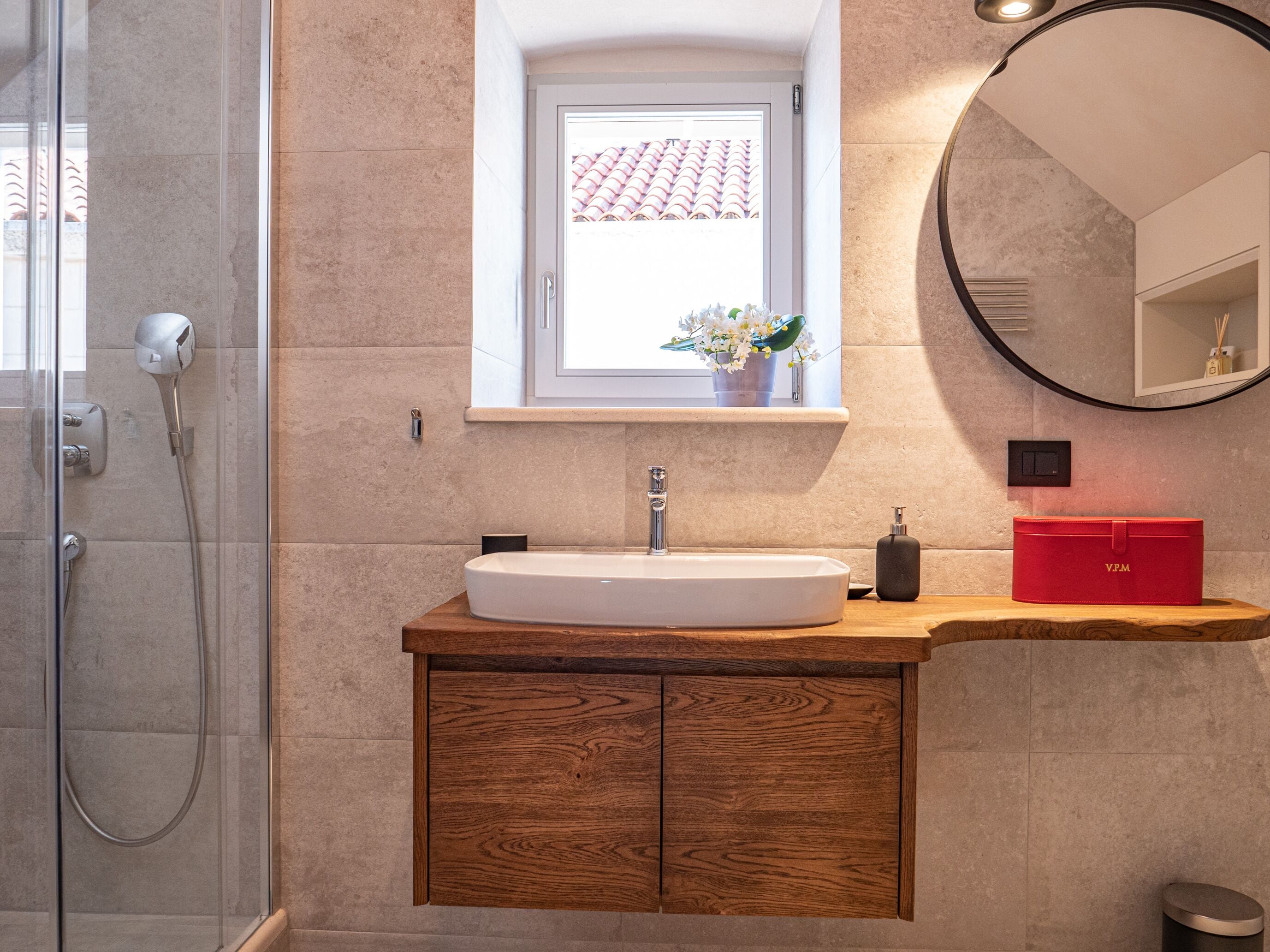 Un baño moderno con cabina de ducha de vidrio, un lavamanos blanco, un mueble de madera, una ventana pequeña con vista a un tejado de azulejos y un espejo redondo.