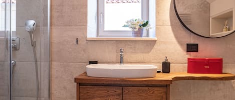 Un baño moderno con cabina de ducha de vidrio, un lavamanos blanco, un mueble de madera, una ventana pequeña con vista a un tejado de azulejos y un espejo redondo.