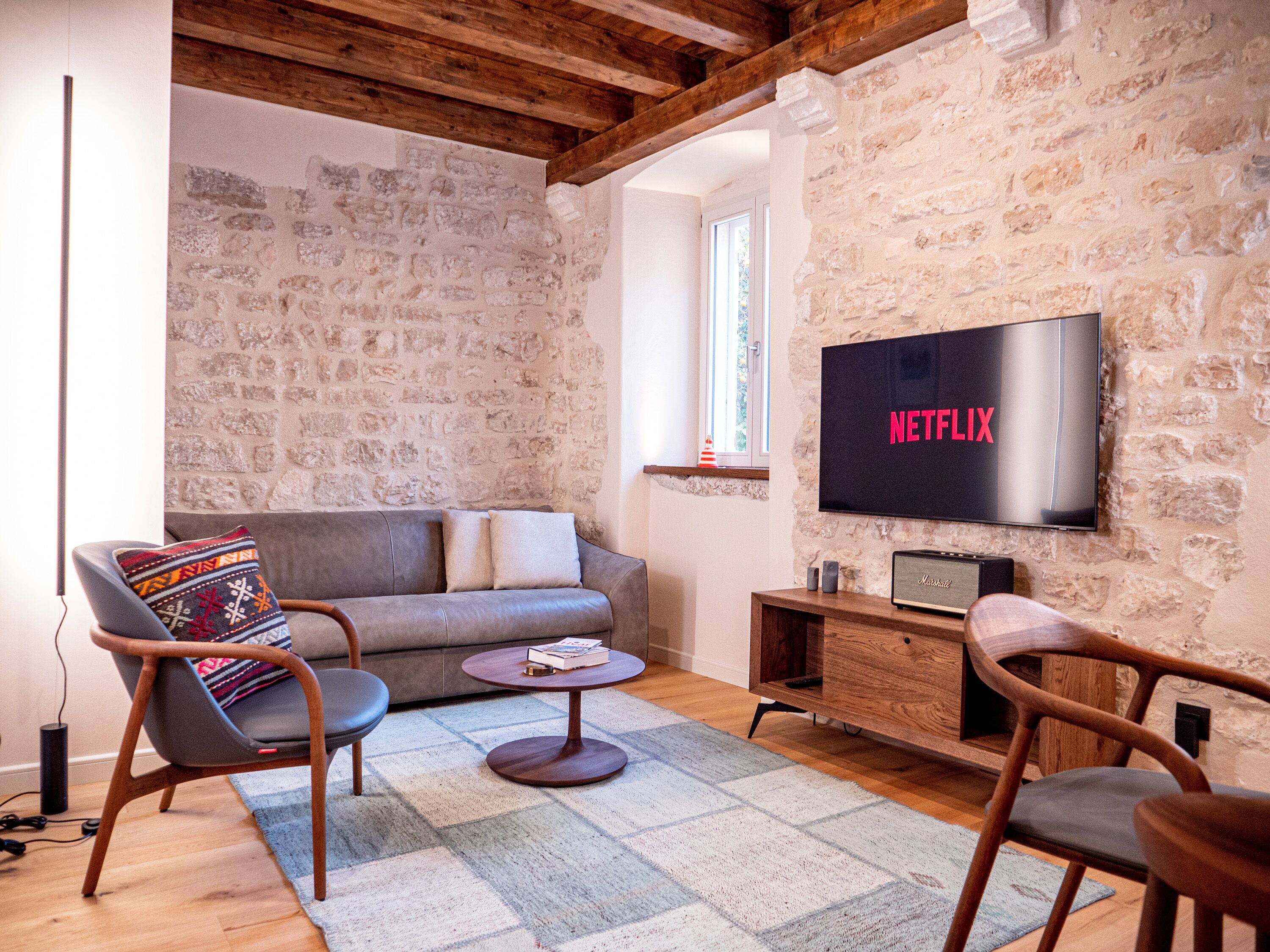 A cozy living room with a stone wall, a grey sofa, a wooden coffee table, and a flat-screen TV mounted on the wall.