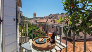 A balcony with a table set for two, featuring a bottle of wine, a glass, and a croissant. A tree with green leaves and an orange fruit is visible in the foreground. - Villa Porta Maestra (Hvar)