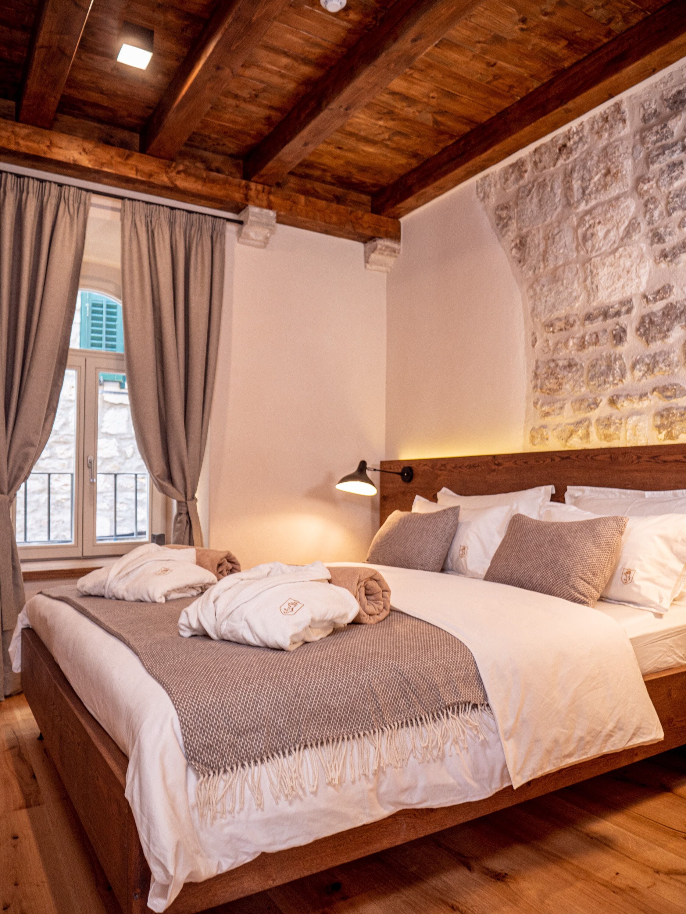 A wooden-framed bedroom with a stone wall, a large bed with white linens, and a window with curtains.