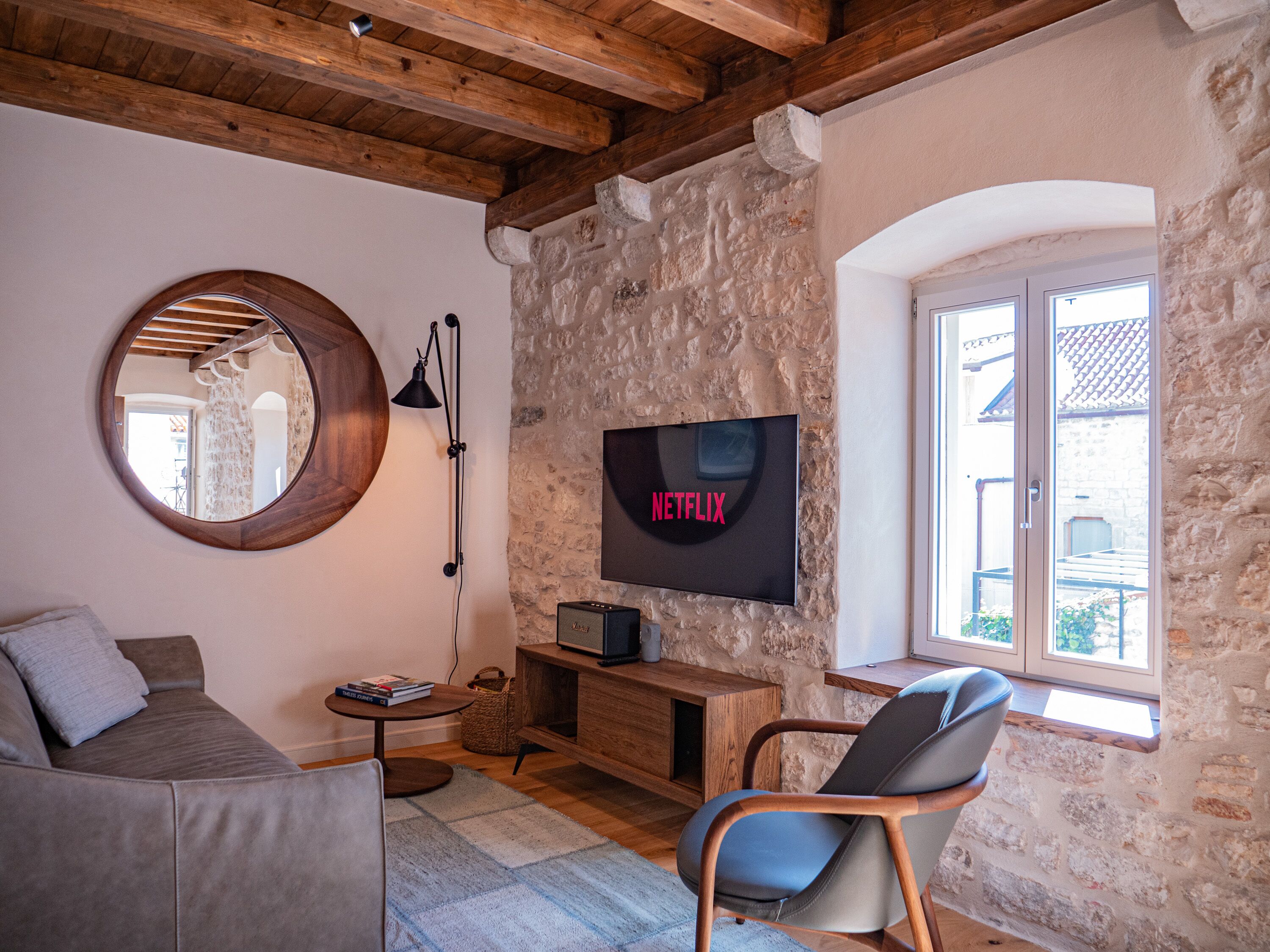 A cozy living room with a stone wall, a round mirror, a wooden ceiling, a TV mounted on the wall, a comfortable sofa, a side table, and a window with a view.