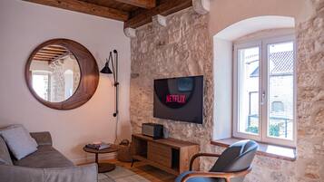 A cozy living room with a stone wall, a round mirror, a wooden ceiling, a TV mounted on the wall, a comfortable sofa, a side table, and a window with a view.