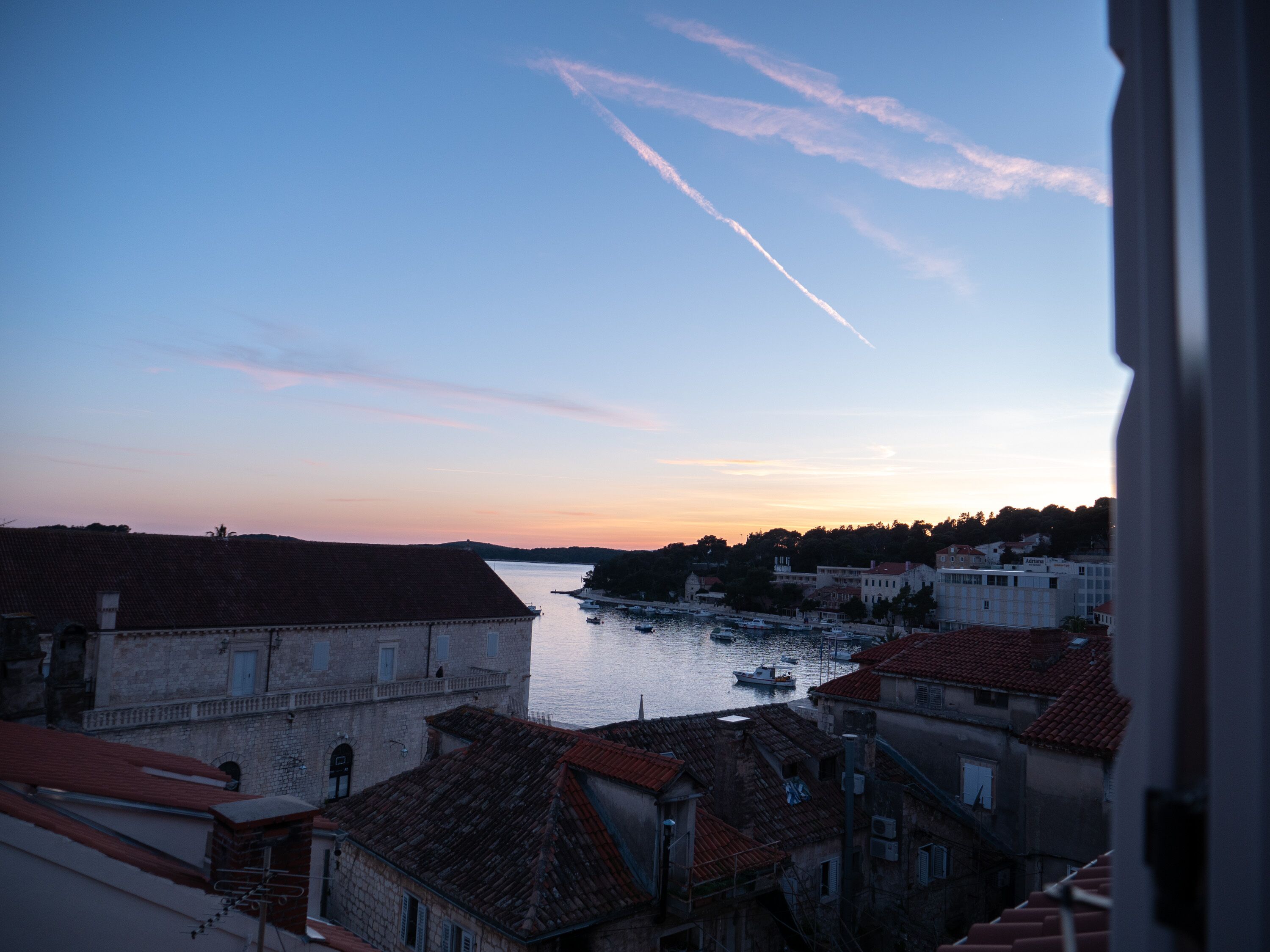 A view of a river with boats, buildings with red roofs, and a clear sky with contrails.