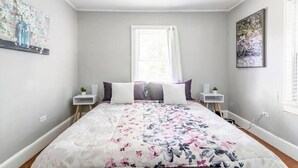 A bedroom with a floral bedspread, a window with white curtains, and artwork on the walls. - {Sunflower Morning} A cozy farmhouse-style house in South End HOT TUB! (Charlotte)