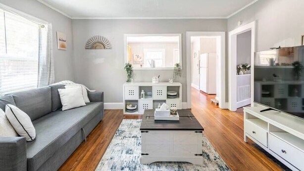 A modern living room with a grey sofa, a coffee table, and a TV stand. There is a white cabinet with decorative items and a mirror above it. - {Sunflower Morning} A cozy farmhouse-style house in South End HOT TUB! (Charlotte)