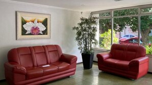 En röd lädersoffa och en fåtölj i en lobby med ett stort fönster, en inramad blomstermålning och en krukväxt med bambu.