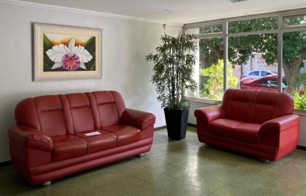 A red leather sofa and chair in a lobby with a large window, a framed flower painting, and a potted bamboo plant.