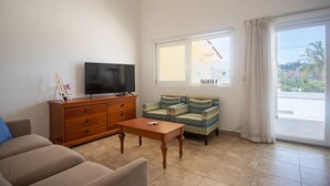 Ein Wohnzimmer mit einer Couch, einem Holztisch, einem Fernseher auf einer Holzkommode und einem Fenster mit Vorhängen.