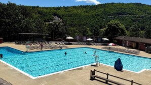 A rectangular swimming pool with a diving board, surrounded by lounge chairs and umbrellas, set against a backdrop of a forested hillside. - Cottage near water park, golf and skiing at Massanutten Resort! (McGaheysville)