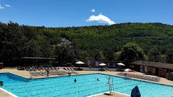 Une piscine rectangulaire dotée d’un plongeoir, entourée de chaises longues et de parasols, le tout situé devant un paysage de colline boisée.