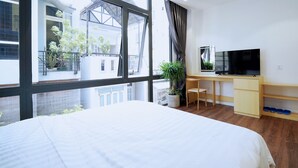 Un dormitorio con cama, escritorio, televisor y ventana con vistas a edificios y plantas.