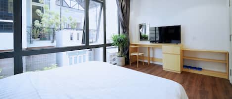 Un dormitorio con cama, escritorio, televisor y ventana con vistas a edificios y plantas.