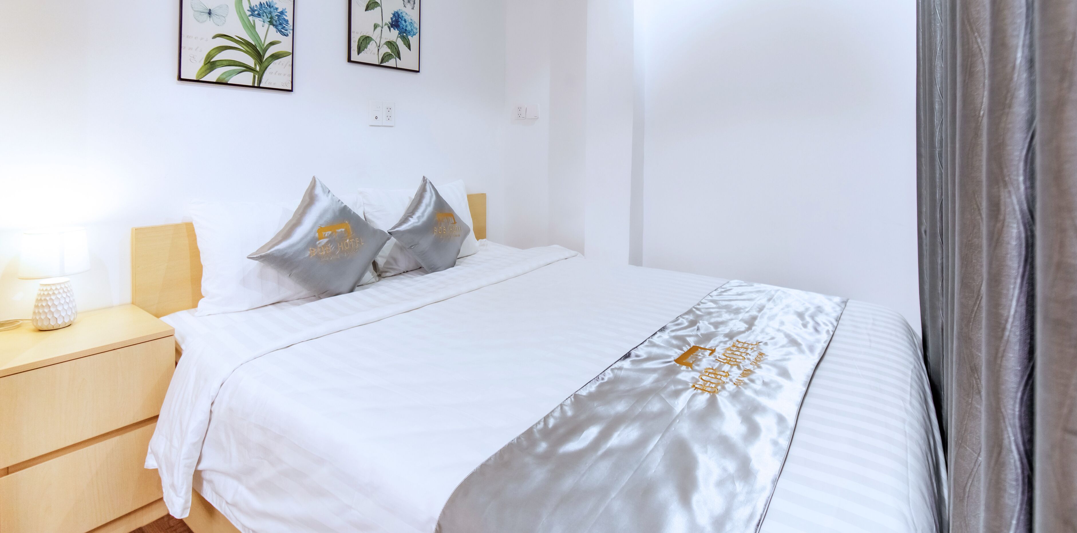 Ein ordentlich bezogenes Bett mit weißer Decke und Kissen, ein Nachttisch mit Lampe sowie zwei gerahmte Pflanzenbilder an der Wand.