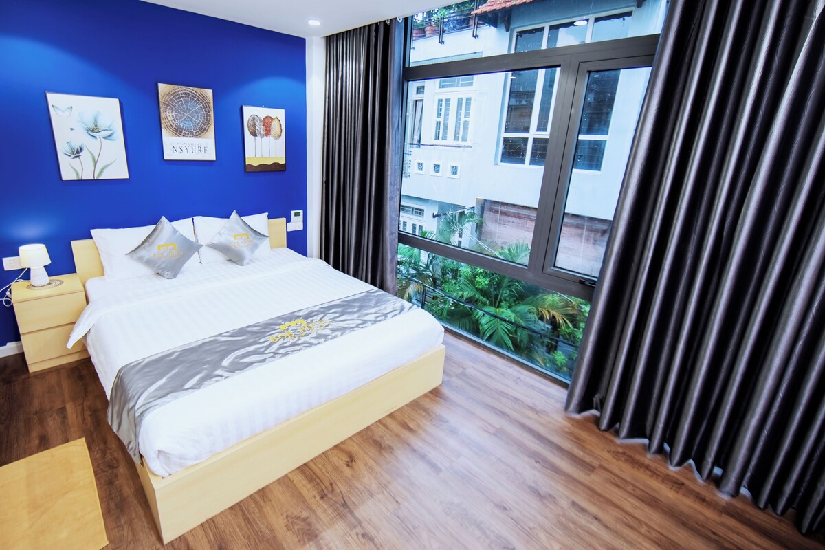 A hotel room with a large bed, bedside tables, and a view of greenery outside.