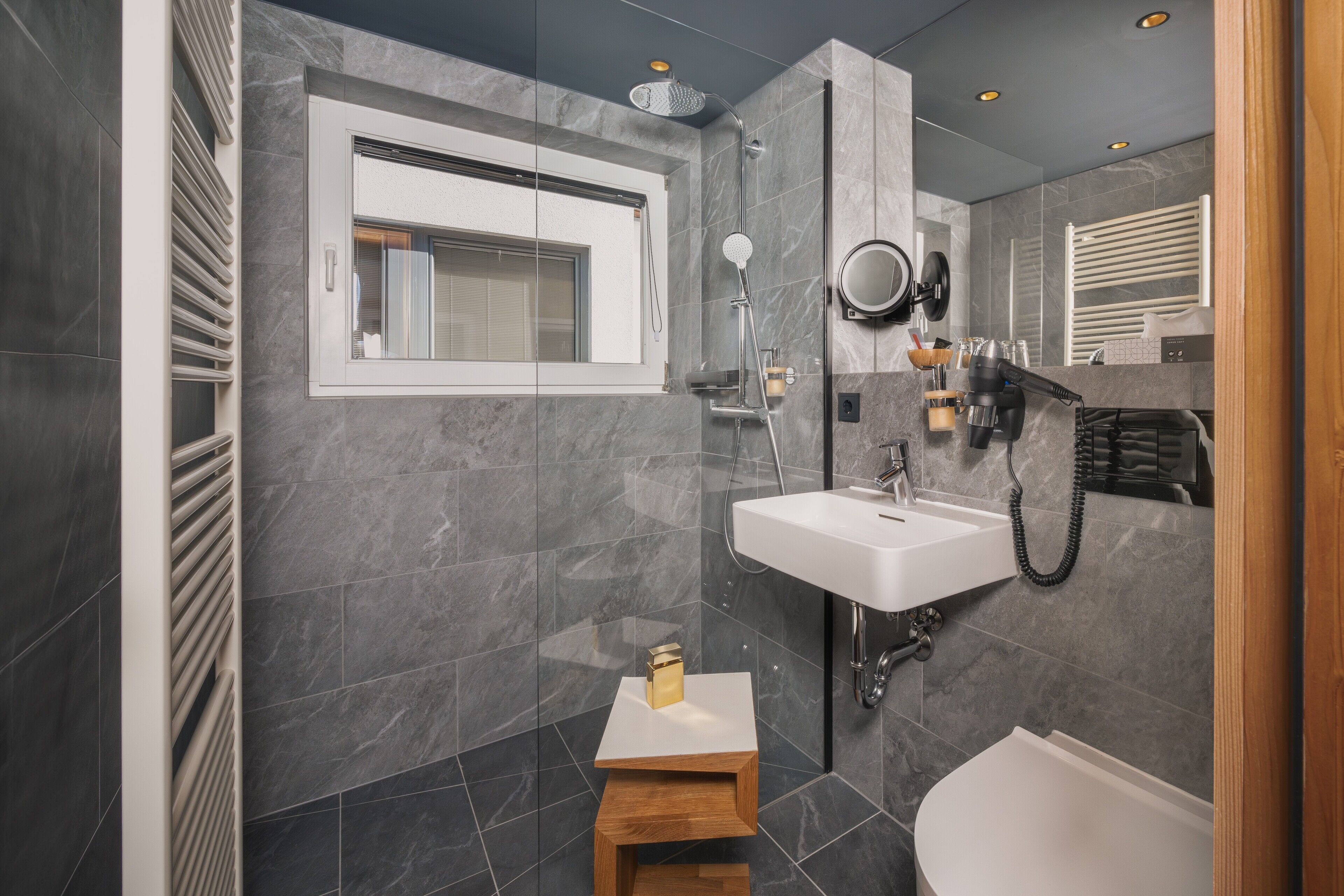 A modern bathroom with a large mirror, a sink, a shower, and a small wooden stool.