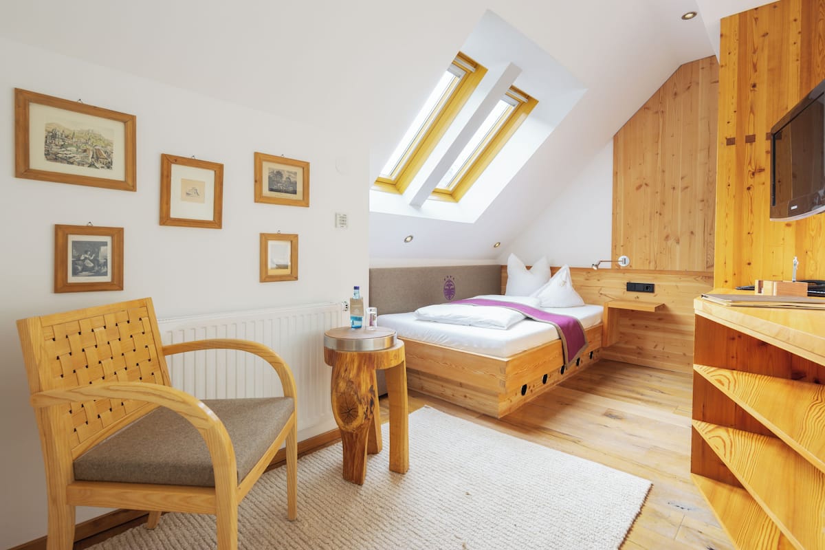 Una habitación con una cama, una silla de madera, una mesita redonda y cuadros en la pared.