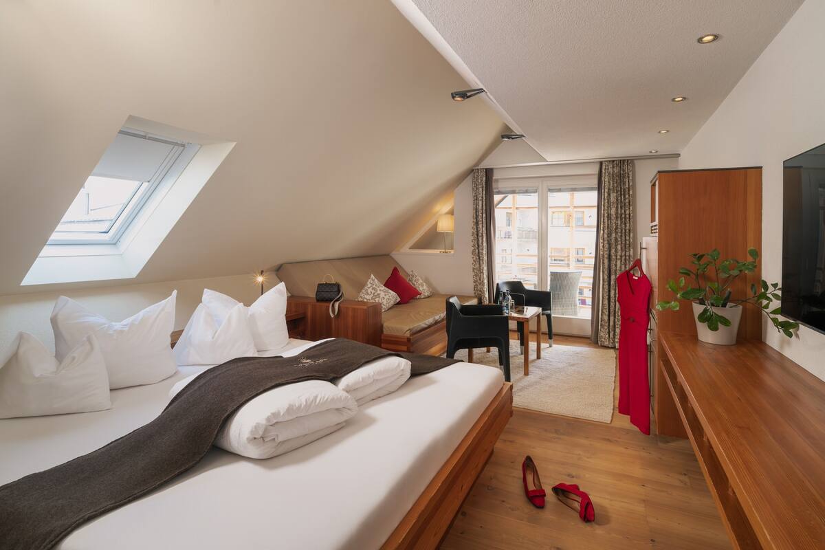 Habitación de hotel moderna con techo inclinado, una cama grande con sábanas blancas, un escritorio con silla, una planta en maceta y vistas por la ventana.