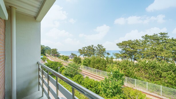 A balcony with a view of a beach, railway tracks, and dense greenery. - Gangneng Badapunggeong Pension (Gangneung)