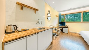 A compact kitchen with a wooden countertop, a kettle, and a sink. There is a small dining area with a chair and a desk with a computer. A window provides a view of trees outside. - Gangneng Badapunggeong Pension (Gangneung)
