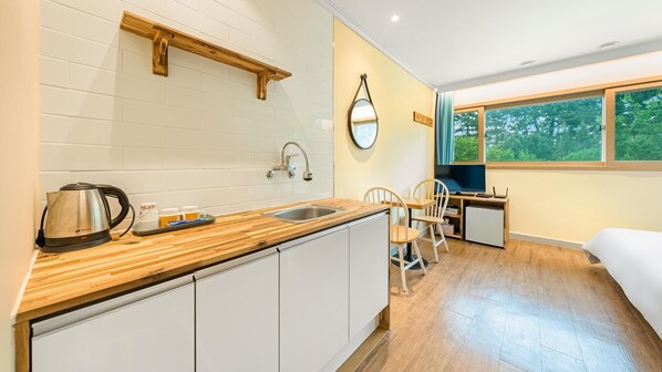 A compact kitchen with a wooden countertop, a kettle, and a sink. There is a small dining area with a chair and a desk with a computer. A window provides a view of trees outside. - Gangneng Badapunggeong Pension (Gangneung)