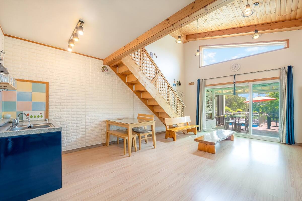 A spacious living area with a wooden staircase, a kitchen with a white brick backsplash, and a large sliding glass door leading to an outdoor patio.
