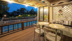 Eine Holzterrasse mit Geländer, die auf einen Pool und Bäume blickt. Zwei Tische und Stühle stehen bereit, und ein Zierzaun mit einem Schild ist zu sehen.
