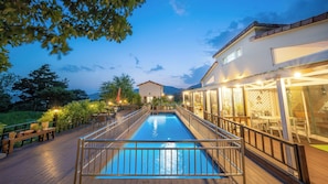 A swimming pool with a wooden deck and metal railing, surrounded by outdoor seating and a building with large windows. - Ganghwado Elinhuvert Pension (Incheon)