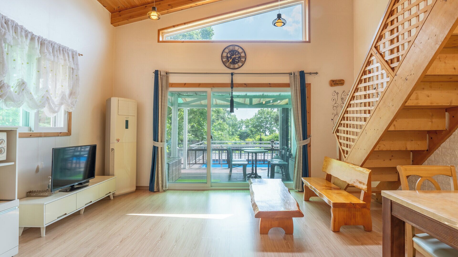 A room with a television, a bench, a table, a staircase, and a view of the outdoors.