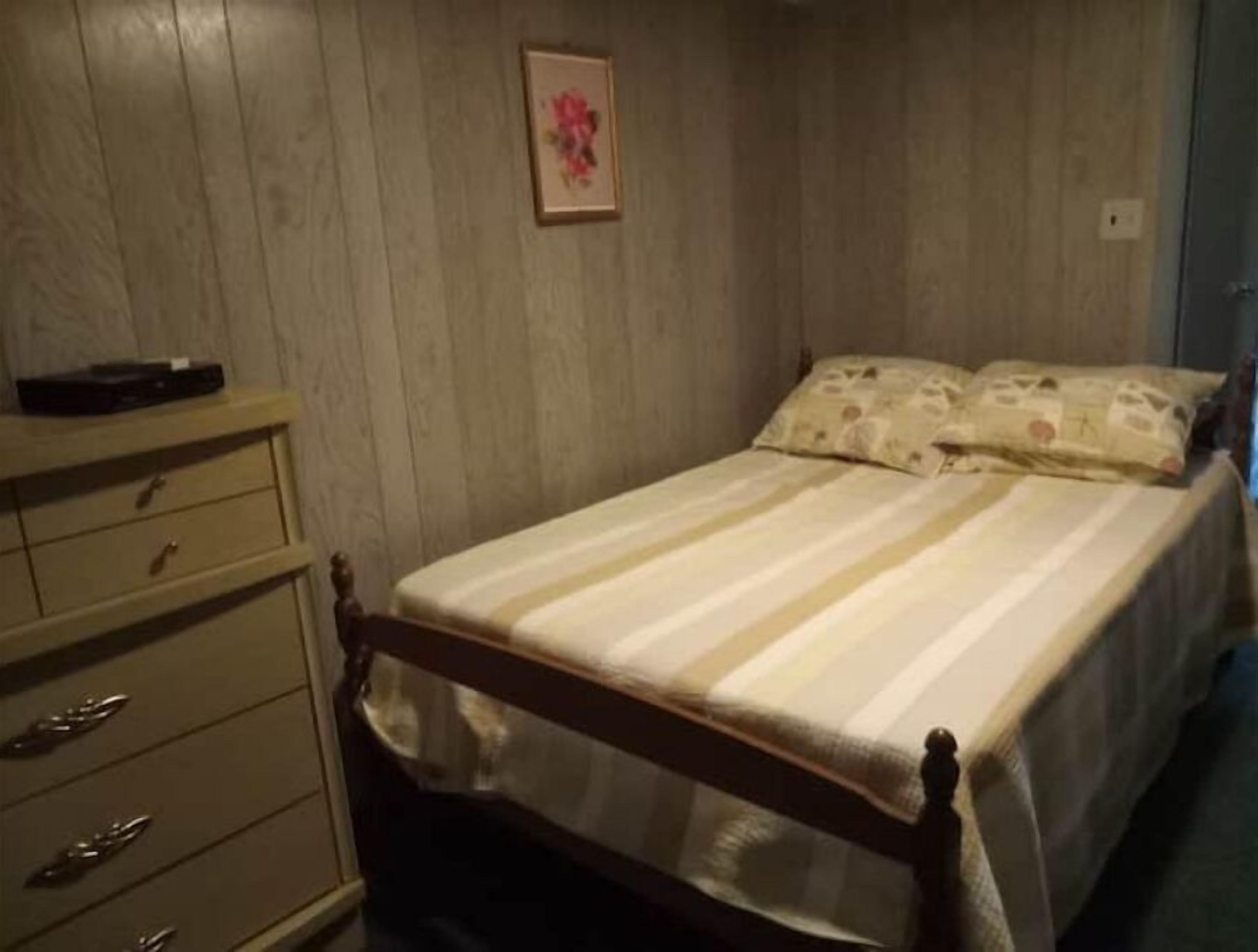 Een slaapkamer met een houten bed, een dressoir met lades en een ingelijst schilderij aan de muur.
