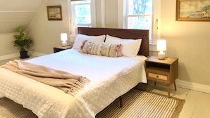 A bedroom with a large bed, two bedside tables, a nightstand, a potted plant, and two windows with curtains. - Quaint & Cozy 1830s Cape- Central Location! (Wiscasset)