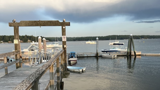 A wooden pier with boats docked at the end, a no swimming sign, and a calm water body with a forested shoreline in the background. - Quaint & Cozy 1830s Cape- Central Location! (Wiscasset)
