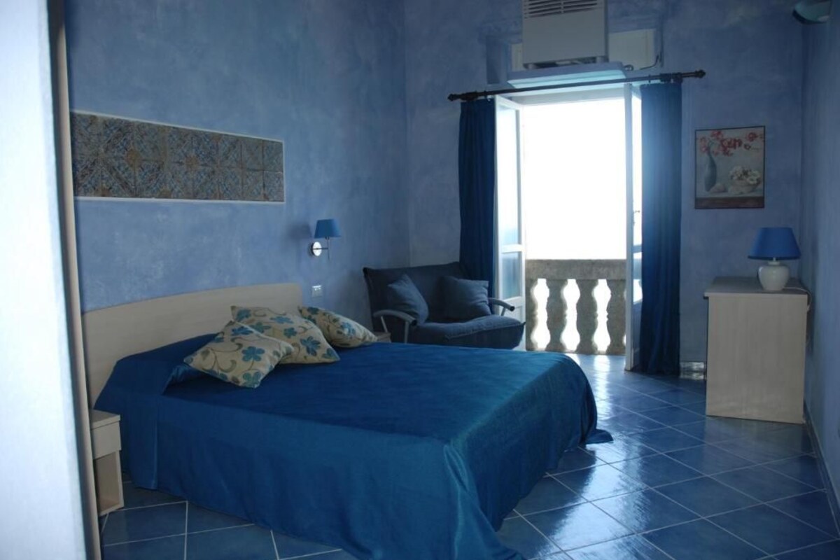 Quarto com cama, cadeira, mesa de cabeceira, luminária e janela com cortinas.