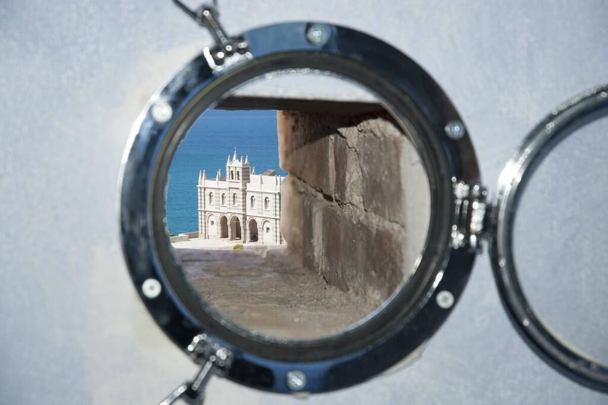 A porthole window with a view of a large, historic building with multiple arches and a clear blue sky in the background.
