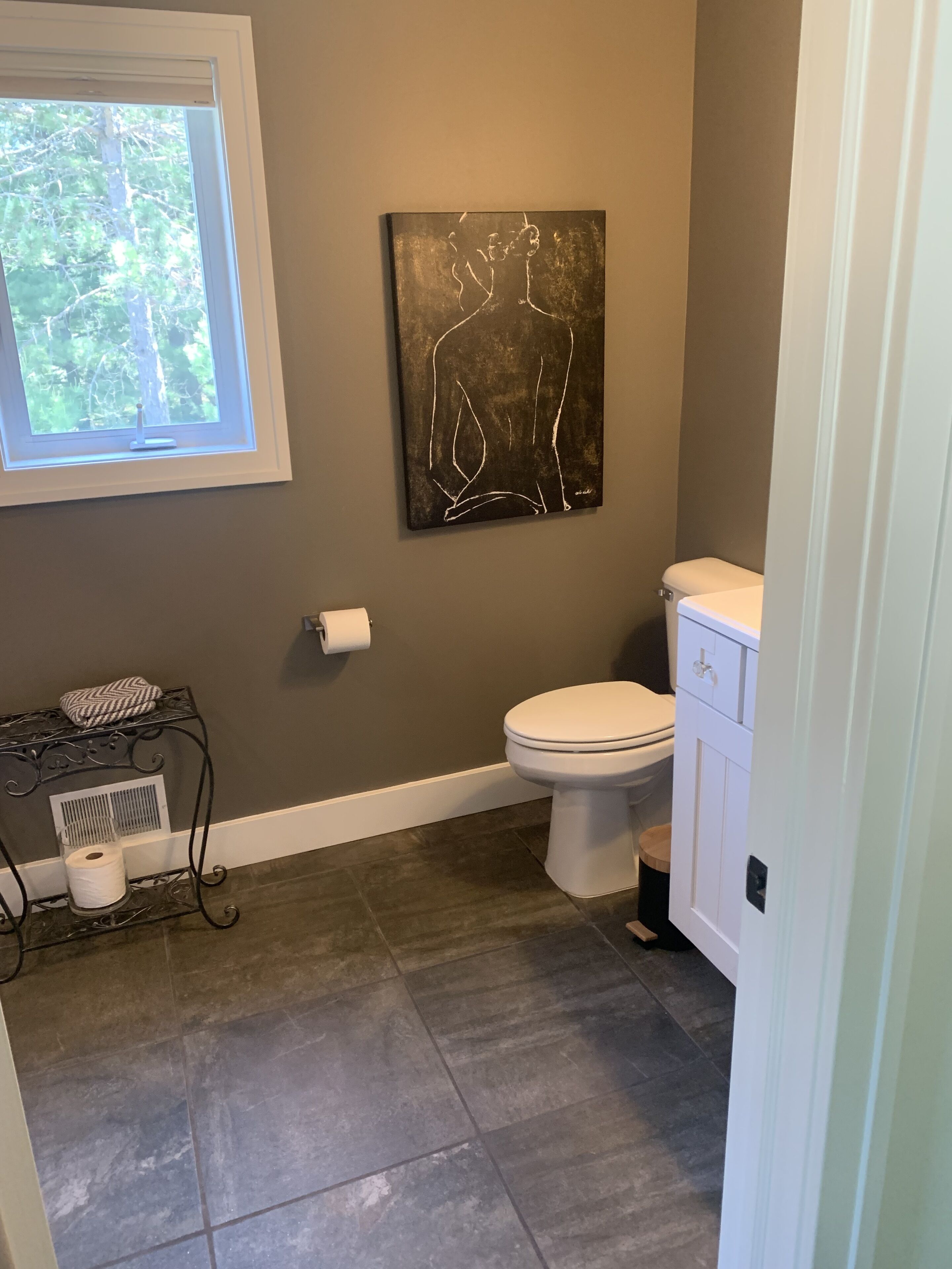 Ein Badezimmer mit Toilette, einem weißen Schrank, einem Handtuchhalter mit Handtuch und Taschentuchbox, einem Lüftungsgitter und einem Fenster mit Blick auf Bäume.