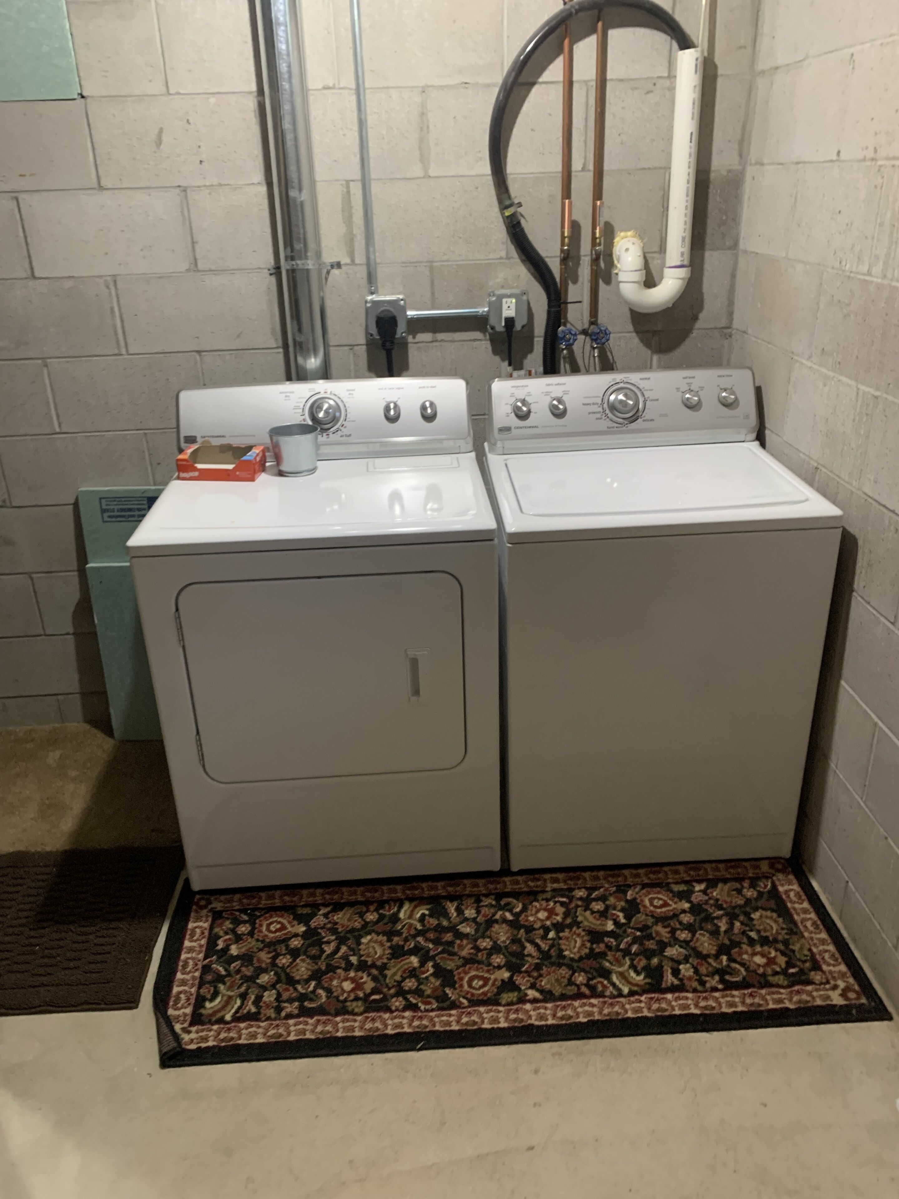 A laundry area with two white front-loading washing machines, a metal pipe, and a power outlet on the wall.