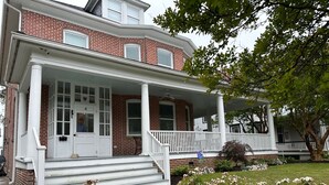 A two-story brick house with a white porch and steps leading to the front door. - Spacious restored 1914 historic home steps from the beach. (Cape Charles)