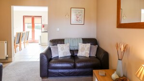 Ein Wohnzimmer mit einem dunkelbraunen Ledersofa, einem Holztisch und einem Spiegel an der Wand. Darüber hängt ein gerahmtes Blumenbild, und eine Tür führt zur Küche.