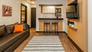 Un espace de vie compact comprenant un coin cuisine, un canapé avec des coussins, un tapis et un téléviseur à écran plat fixé au mur.