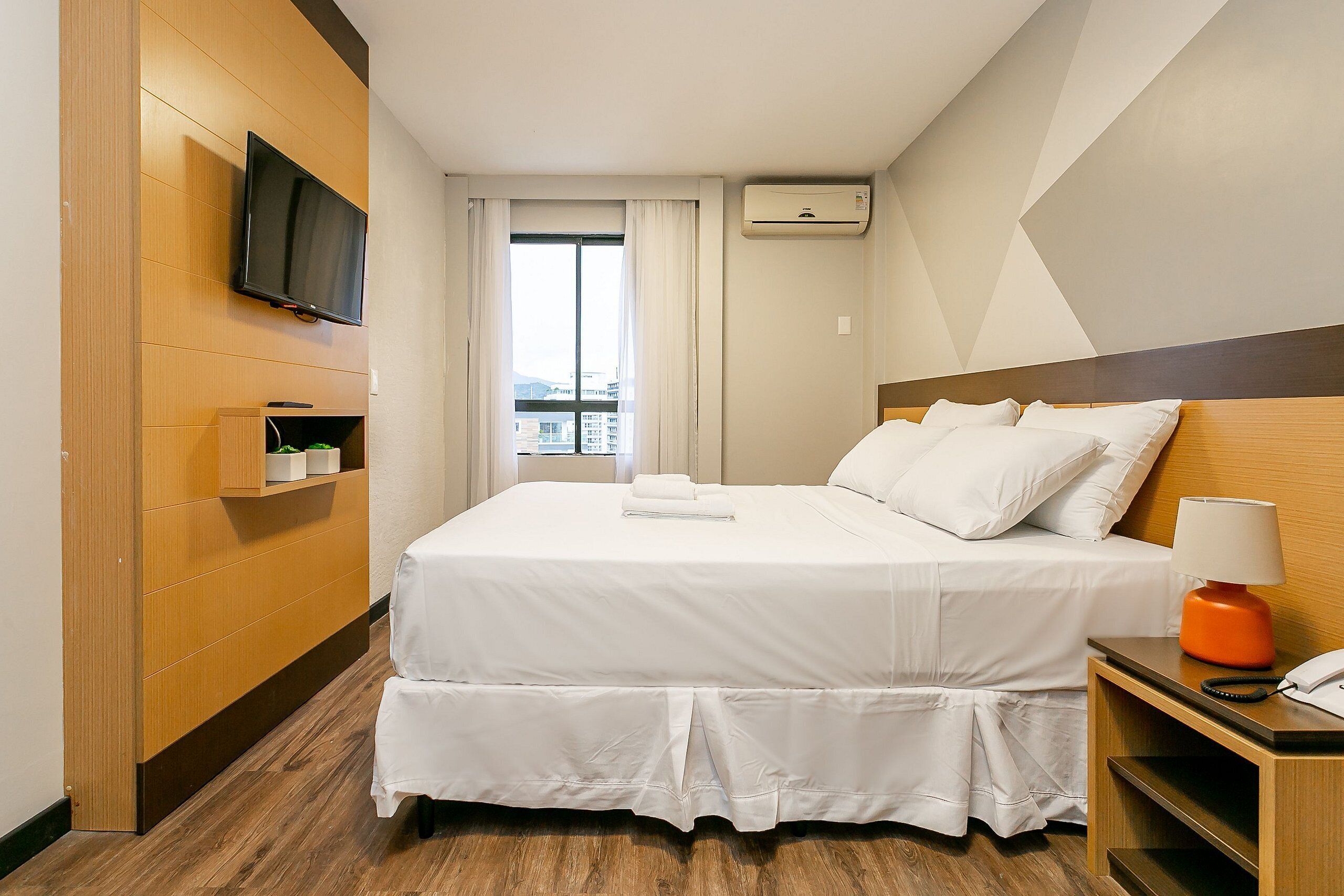 Una habitación de hotel con una cama, un televisor montado en la pared, una mesita de noche con lámpara y una ventana con cortinas.