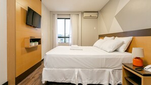 Une chambre d’hôtel avec un lit, un téléviseur fixé au mur, une table de chevet avec une lampe et une fenêtre avec des rideaux.