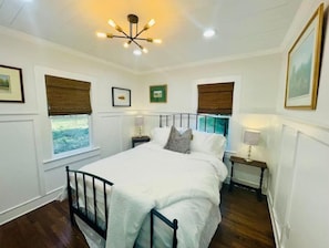 Une chambre à coucher comprenant un lit, deux tables de chevet, un luminaire au plafond et des fenêtres équipées de stores.