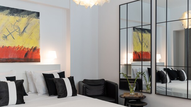 Una habitación de hotel moderna con una cama grande, almohadas blancas y negras, una mesita con un florero y un espejo grande que refleja la habitación.