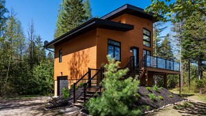 A modern wooden cabin with a staircase leading to a deck, surrounded by trees.