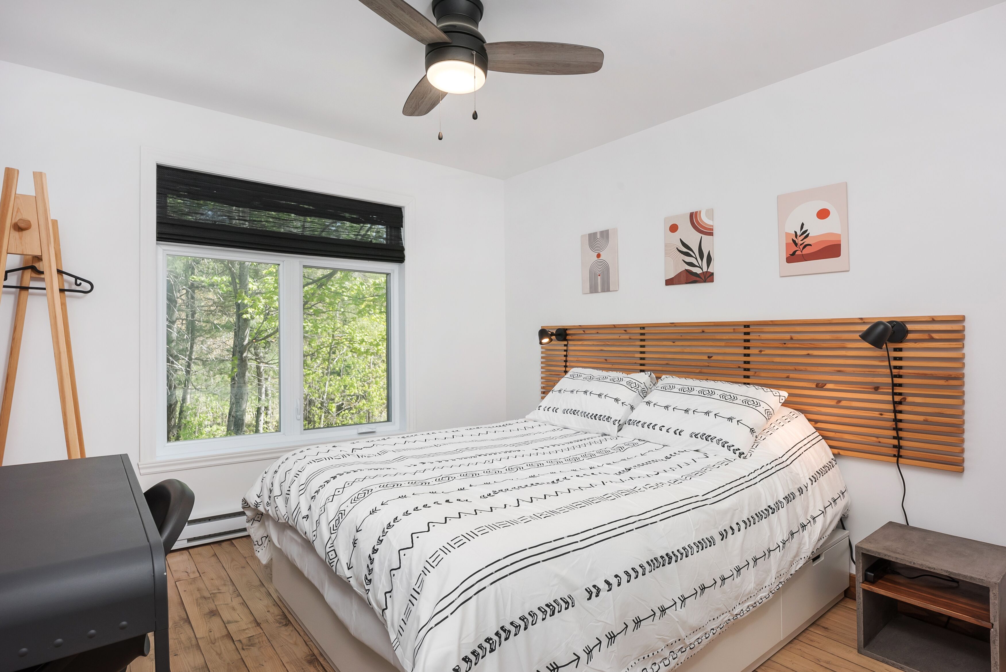 Ein Schlafzimmer mit Bett, Schreibtisch, Stuhl, Deckenventilator, Fenster mit Jalousien und Kunstwerken an der Wand.