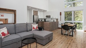 A modern living room with a gray sectional sofa, a small round table, and a dining area with a wooden table and chairs. There is a kitchen area with white cabinets and a refrigerator. Large windows provide natural light and a view of trees.