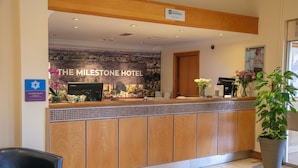 A reception desk at The Milestone Hotel with a cityscape backdrop, a "Save Hand Promoter" sign, and a potted plant. - The Milestone Hotel, Peterborough (Cambridgeshire), Sure Collection by Best Western (Peterborough)