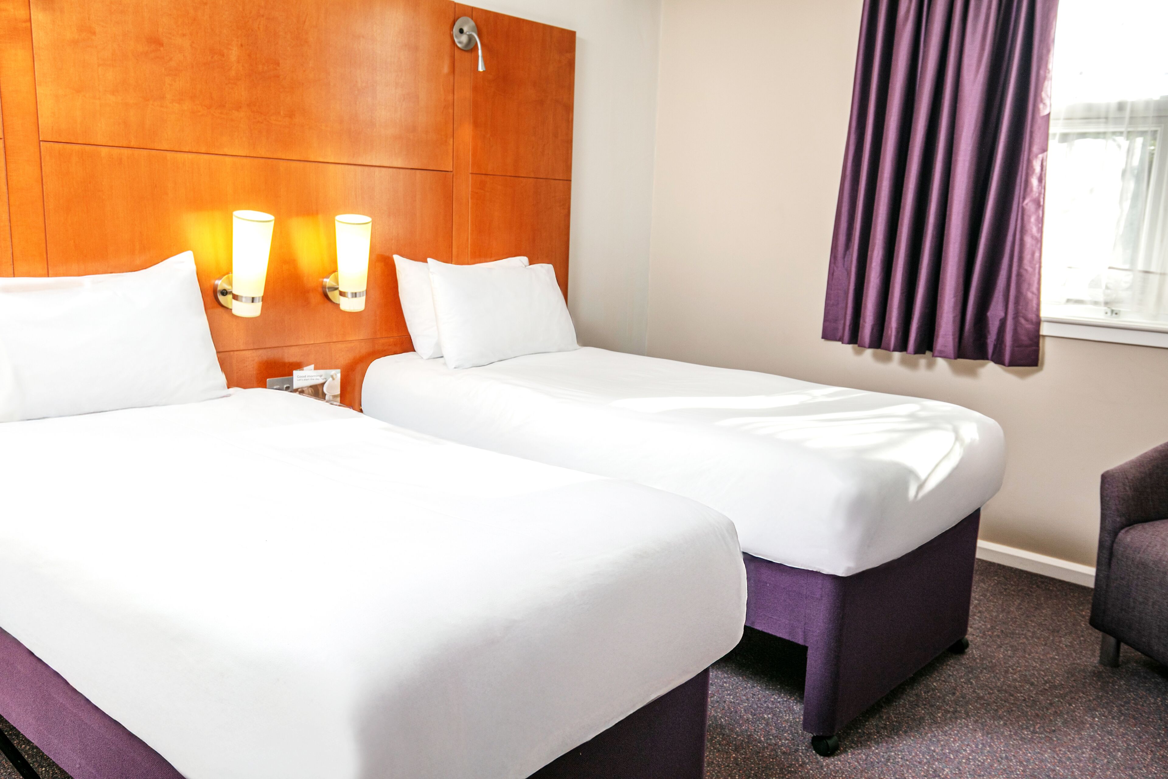 A hotel room with two beds, a wooden headboard, wall-mounted lights, a purple chair, and a window with purple curtains.