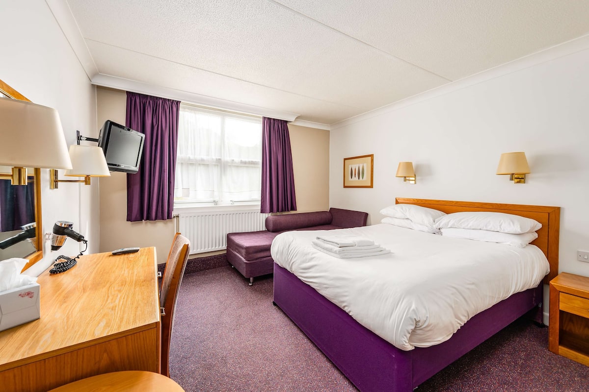 A hotel room with a bed, a desk with a computer, a chair, a small table, a TV, and a window with curtains.
